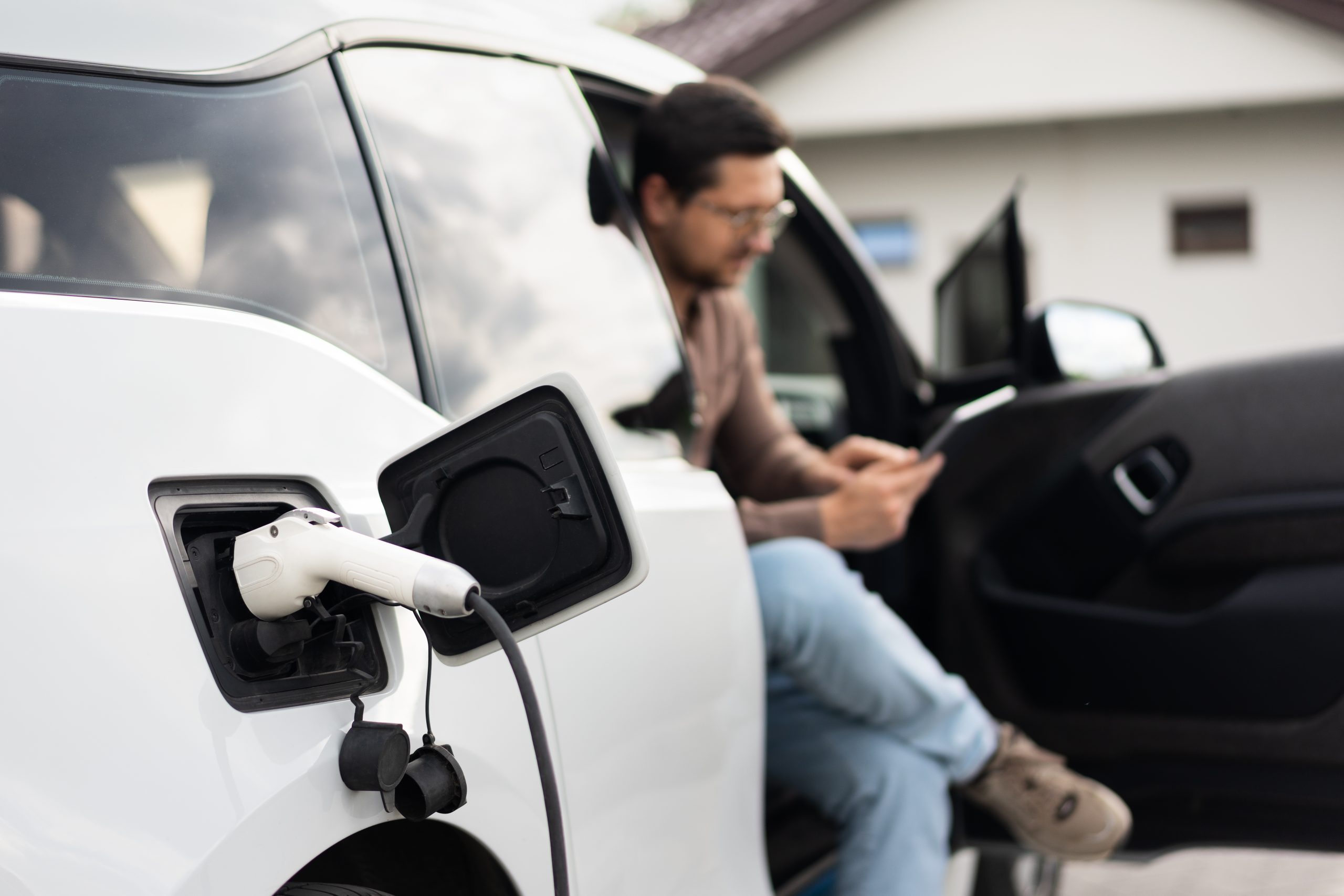 Electric Car Charging While Man Uses Smartphone, Eco-Friendly Transportation And Technology Concept.